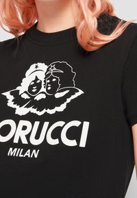 Gros plan sur un t-shirt noir avec deux visages d'anges blancs jumeaux et le texte "ORUCCI MILAN" imprimé sur la poitrine.
