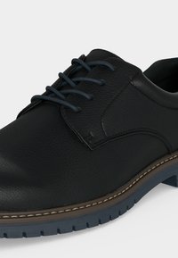 Chaussure habillée pour homme en cuir noir à lacets, dessus texturé, intercalaire marron cousue et semelle extérieure robuste en caoutchouc bleu foncé.