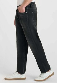 Jean noir en denim avec une coupe droite, cinq poches et des coutures visibles. Portés avec des baskets blanches à semelle marron.