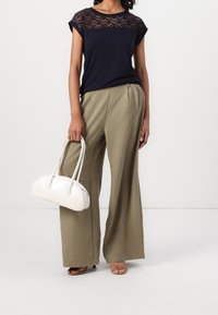 Námořnický top s krajkovým hrdlem, volné široké kalhoty olivové barvy a bílá taška s zakřiveným designem a dvěma rukojeťmi; sandály na otevřenou špičku.
