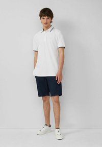 Weißes Poloshirt mit marineblauen Akzenten, kombiniert mit dunkelblauen Shorts. Das Model trägt weiße Sneakers. Einfarbiger Hintergrund.