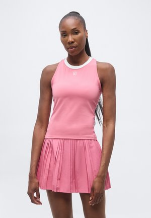 Femeie purtând un top sport roz fără mâneci și o fustă plisată roz, stând în fața unui fundal simplu și luminos.