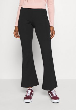 Femme portant un pantalon évasé noir, un haut à manches longues rose clair et des baskets rouges et blanches, debout devant un fond uni.