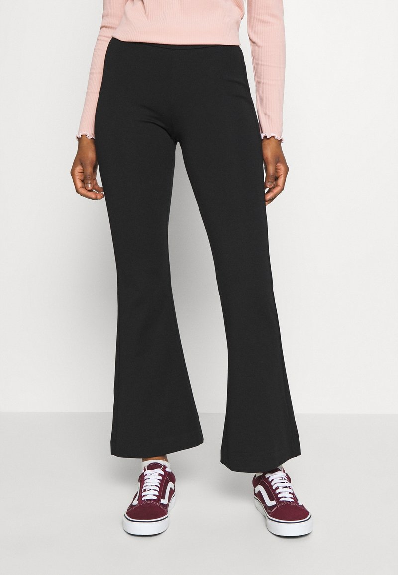 Femme portant un pantalon évasé noir, un haut à manches longues rose clair et des baskets rouges et blanches, debout devant un fond uni.