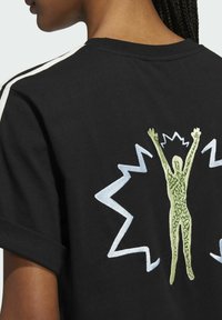 T-shirt in cotone nero con una grande figura ricamata verde con le braccia sollevate, circondata da accenti blu a zigzag sul retro.