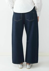 Osoba w ciemnoniebieskich szerokich dżinsach i srebrnych trampkach, zwrócona tyłem, z białą koszulą widoczną z tyłu.