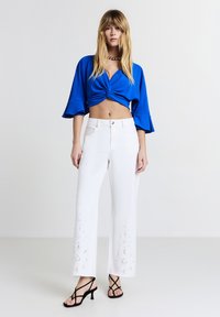 Top blu cropped con frontale attorcigliato, pantaloni bianchi a gamba larga con motivi floreali ricamati sull'orlo e sandali neri con tacco e cinturini.