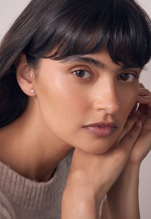 Junge Frau mit braunen Augen und dunklem Haar, die ihr Kinn auf gefaltete Hände stützt, trägt einen beigen Pullover und kleine runde Ohrstecker.