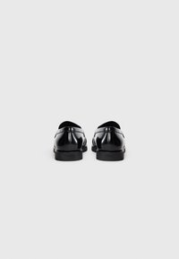 Must musta mustaäsi mustad läikivad nahast loaferid, vaadatud tagant, lihtsalt valge tausta peal.