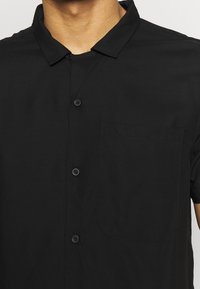 Musta lyhythihainen paita, jossa nappikiinnitys edessä, vasemmalla puolella tasku ja sileä, kevyt kangas.