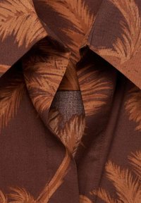 Camicia marrone con motivo di foglie di palma in tonalità di arancione. Presenta un colletto a punta e un tessuto dalla consistenza liscia.