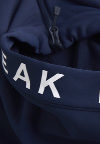 Blå sweatshirt med en stoftekstur, der har et hvidt trykt logo bånd og en lynlåslukning i kraven. Accentueret med en lynlåstræk.