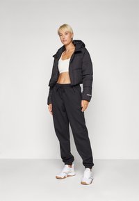 Svart kort puffjakke med hette, hvit tettsittende crop-top, svarte joggebukser og hvite joggesko med gummisåler. Enkelt design og uformell passform.