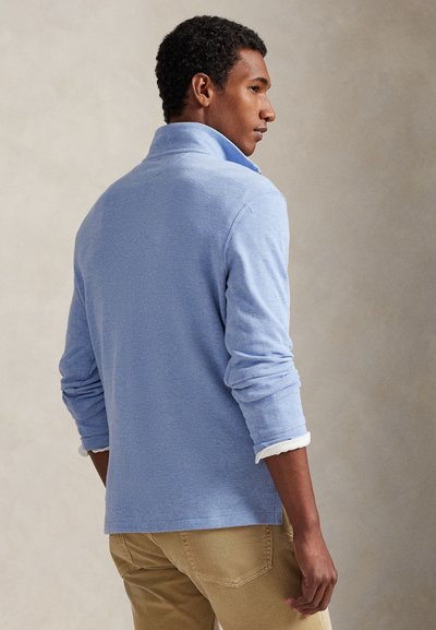 Homme portant une chemise bleu clair à manches longues avec col, pantalon beige, debout, dos partiellement tourné contre un fond neutre.