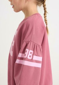 Sweat-shirt rose avec manches bouffantes, arborant les chiffres blancs audacieux "98" et des accents rayés sur les manches. Confectionné en coton doux.