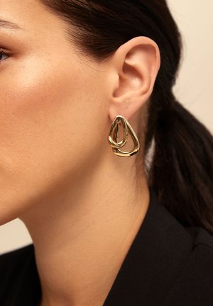 Pendientes de doble aro dorados usados por una mujer con el cabello oscuro recogido, mostrados en primer plano contra un fondo neutro.