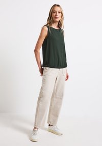 Mörkgrön ärmlös topp med lös passform, i kombination med ljusbeige vida byxor. Modellen bär vita sneakers med texturdetaljer.
