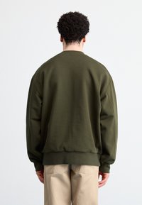 Sudadera verde oliva con un corte relajado, puños y dobladillo acanalados, textura suave y detalles de costura. Se muestra la vista trasera.