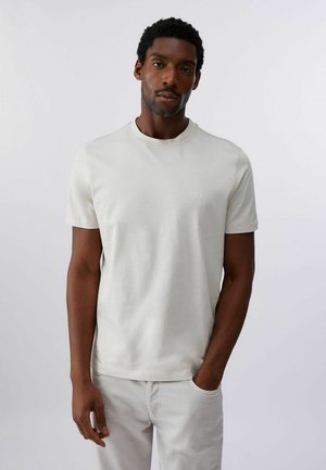 Uomo con capelli corti e ricci che indossa una maglietta beige chiaro tinta unita e pantaloni chiari, in piedi con una mano in tasca.