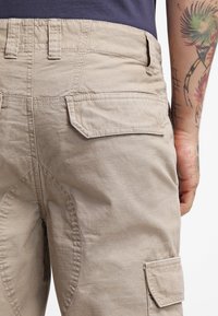 Beige cargo pants with a textured fabric, featuring two patch pockets on the back and visible stitching details. Waistband with belt loops.