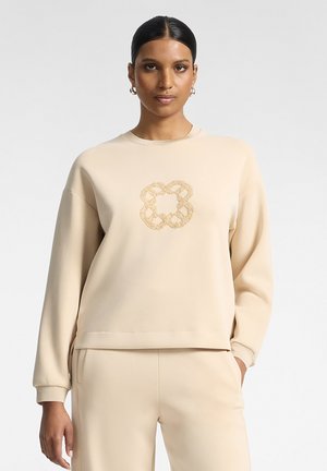 Femme portant un sweat-shirt beige et un pantalon assorti avec un motif de nœud tressé doré sur la poitrine, debout devant un fond clair uni.