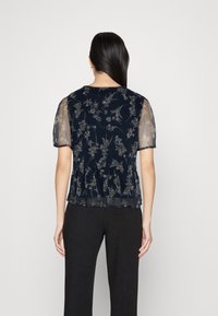 Blusa floreale blu navy con maniche corte trasparenti, caratterizzata da un orlo peplum e motivi botanici intricati in colori chiari. I pantaloni neri sono indossati sotto.