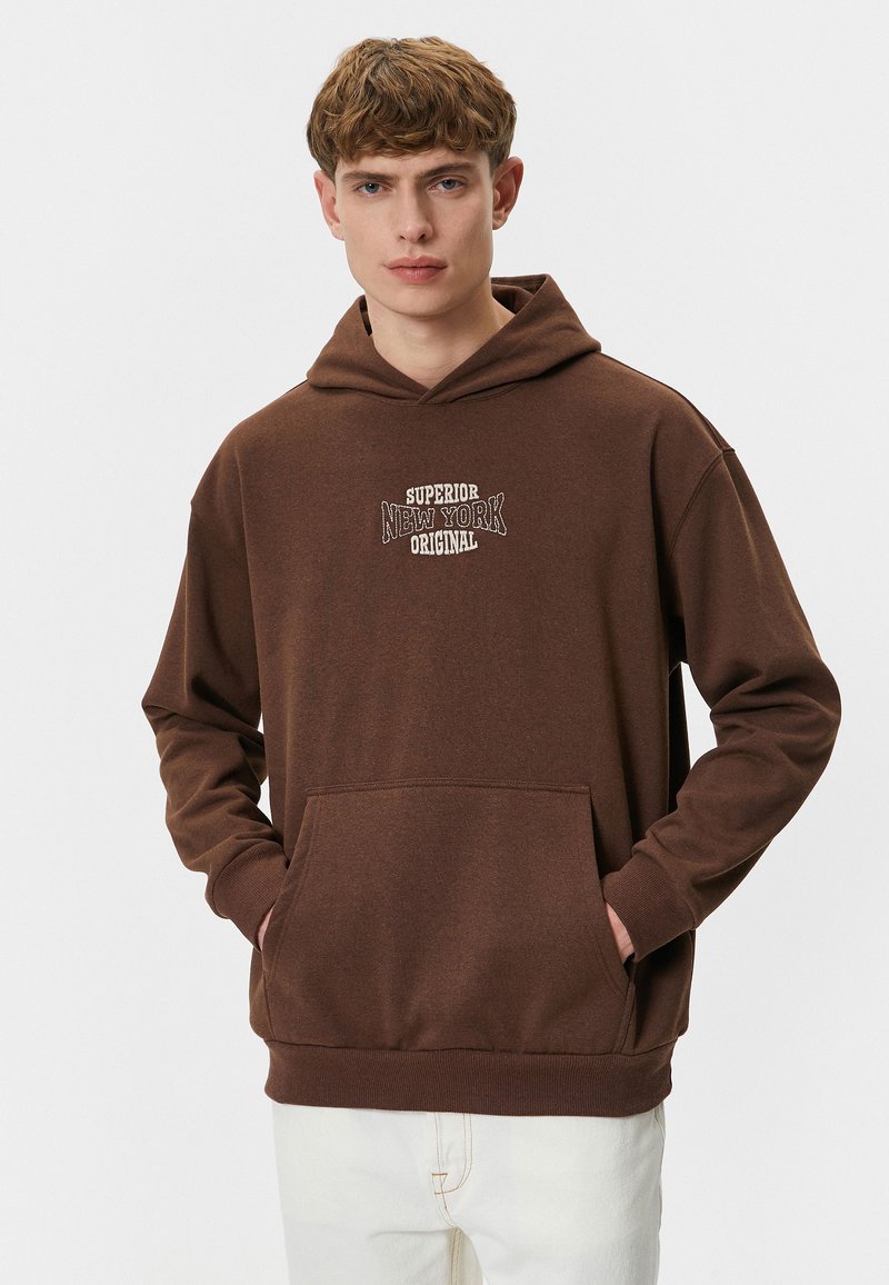 Brun hættejakke med en stor frontlomme og snøre i hætten. Har hvid tekst "Superior New York Original" i en vintage skrifttype.