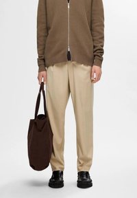 Pantalones beige combinados con un cárdigan de punto marrón con cremallera. El conjunto incluye un bolso tote marrón y zapatos negros con detalles texturizados.