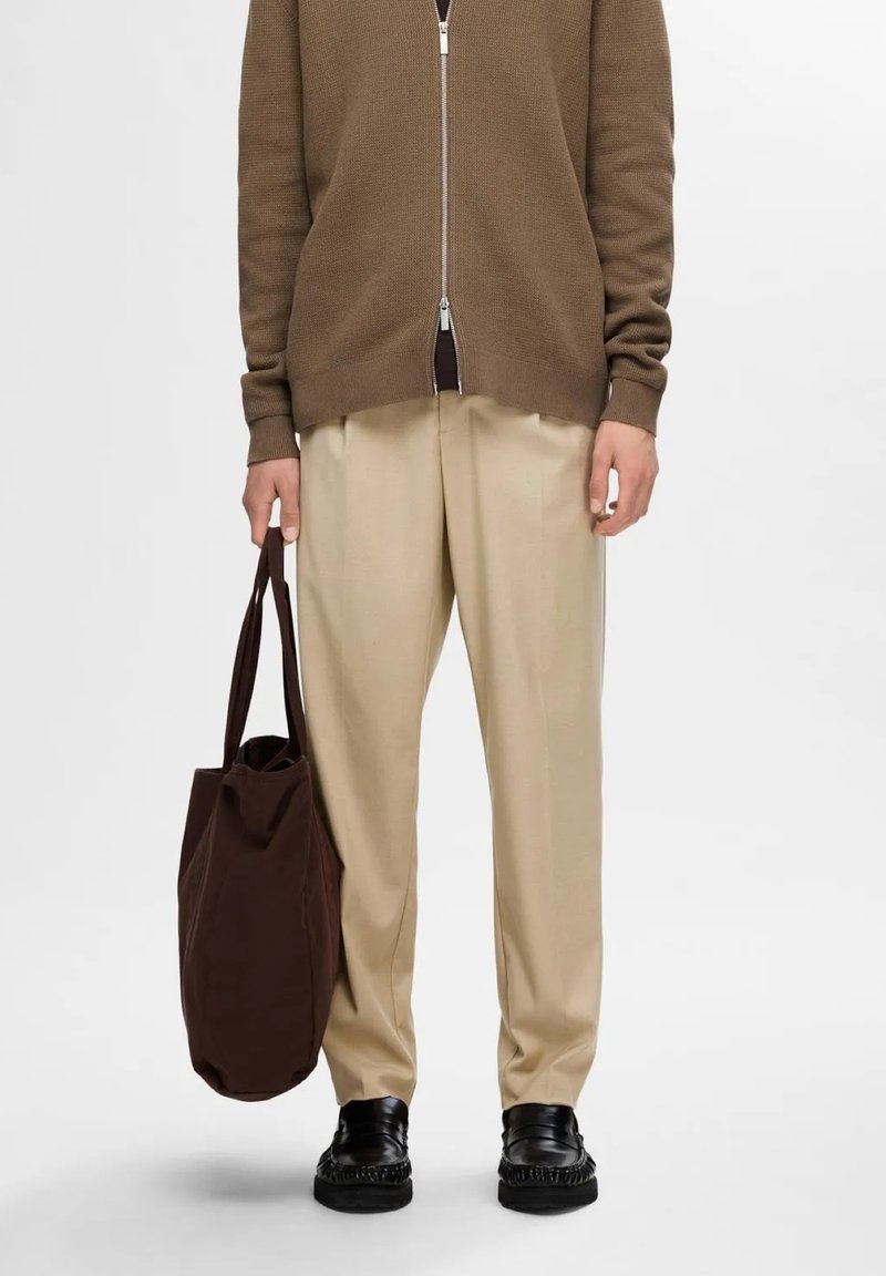 Pantalones beige combinados con un cárdigan de punto marrón con cremallera. El conjunto incluye un bolso tote marrón y zapatos negros con detalles texturizados.