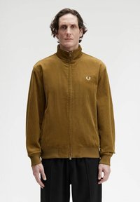 Uomo con capelli ricci corti, che indossa una giacca in velluto a coste marrone con zip e pantaloni neri, in piedi davanti a uno sfondo bianco semplice.