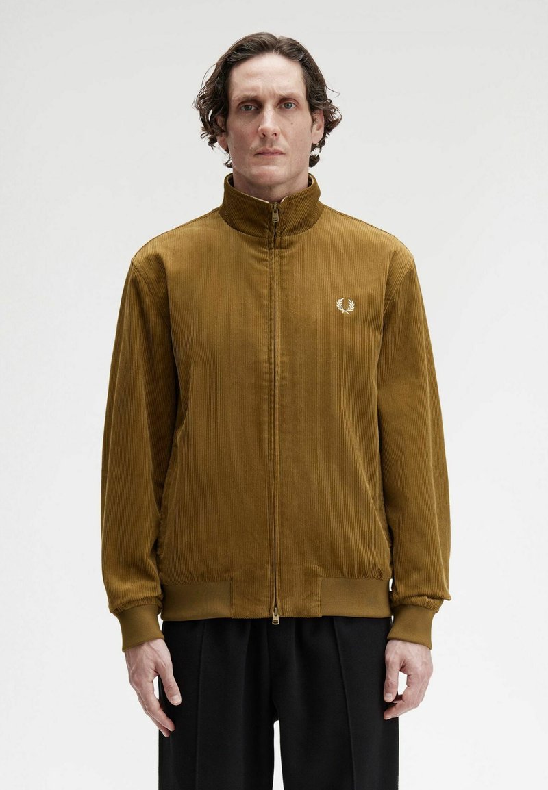 Uomo con capelli ricci corti, che indossa una giacca in velluto a coste marrone con zip e pantaloni neri, in piedi davanti a uno sfondo bianco semplice.