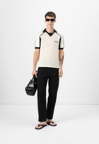 Hvit stripete polo skjorte med svarte detaljer, svarte bukser, svarte flip-flops og en svart veske. Modellen har på seg solbriller. Minimalistisk bakgrunn.