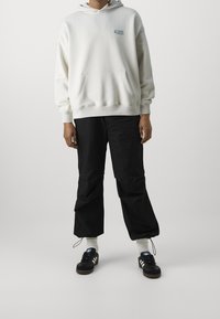 Felpa bianca con cappuccio e tasca frontale, pantaloni neri larghi, calzini bianchi e sneaker nere con tre strisce bianche.