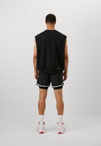 Black sleeveless sweatshirt with a crew neck, paired with black shorts featuring white side stripes. Model wears white and pink sneakers.