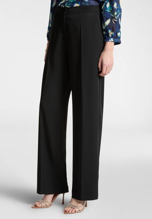 Pantaloni neri a gamba larga con pieghe frontali, design su misura e texture liscia. Abbinati a sandali con tacco alto e cinturini.