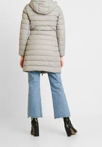 Lichtgrijze pufferjas met een capuchon, geborduurde ontwerp en lange mouwen. Draagt boven op blauwe flared jeans en zwarte enkellaarsjes met een blokhak.