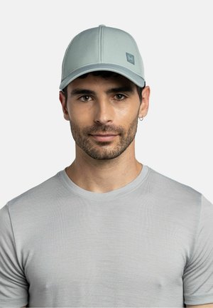 Hombre con gorra de béisbol gris claro y camiseta de cuello redondo a juego en gris claro, con cabello corto y oscuro y pequeño pendiente de aro en la oreja izquierda.