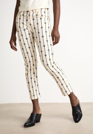 Widok na nogi osoby noszącej kremowe spodnie z pionowymi granatowymi kwiatowymi paskami oraz czarne wsuwane buty na obcasie, stojącej z jedną nogą wysuniętą do przodu.