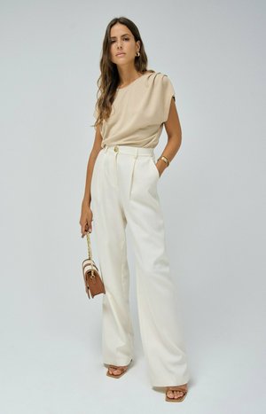 Top drapeado beige, pantalones blancos de cintura alta y bolso marrón con cadena dorada. Modelo llevando sandalias de punta abierta y joyería minimalista de oro.