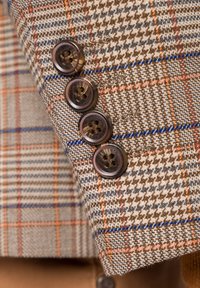 Brązowa tkanina w wzory z pomarańczowymi i niebieskimi akcentami; posiada cztery okrągłe ciemne guziki na mankiecie. Tekstura jest tkana i strukturalna.