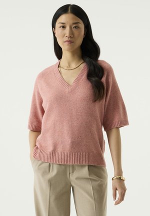 Femme aux longs cheveux foncés portant un pull rose à col en V, un pantalon beige, un bracelet doré et un collier chaîne, debout sur un fond uni.