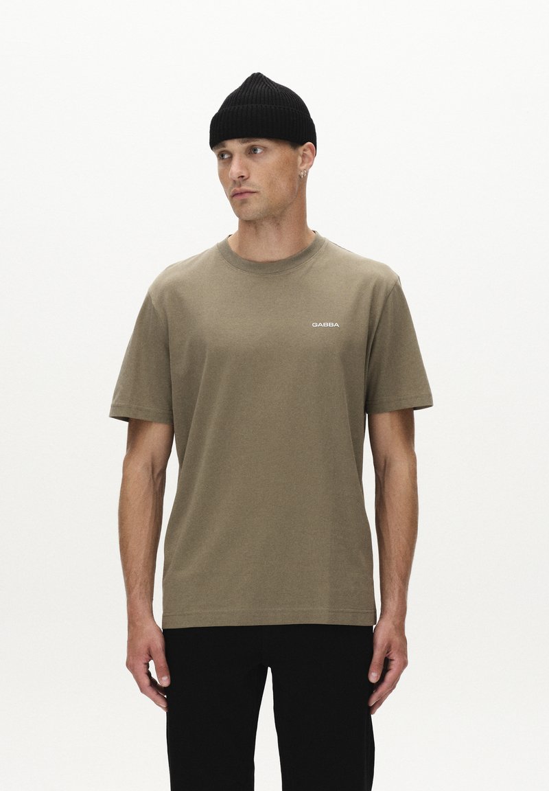 Olijfgroene katoenen t-shirt met korte mouwen, ronde hals en klein "GABBA" logo; gecombineerd met zwarte broek en een zwarte geribbelde beanie.