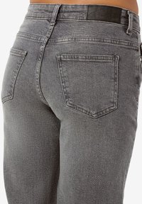 Vedere din spate a unei persoane care poartă blugi gri din denim cu talie înaltă, două buzunare în spate și o etichetă de brand neagră pe talie.