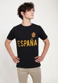 Sort bomulds T-shirt med gult "ESPAÑA" tekst og et farverigt våbenskjold på venstre brystområde. Korte ærmer og rund halsudskæring.