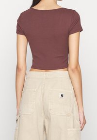 Personne portant un crop top marron ajusté à manches courtes et un pantalon cargo beige avec de grandes poches arrière et un logo de marque visible sur la poche droite.