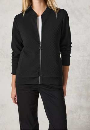Vrouw die een zwart zip-up jack met structuur en geribbelde manchetten draagt, samen met een zwarte broek, staand tegen een lichtgrijze achtergrond.