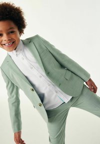 Completo per bambini verde chiaro realizzato in tessuto testurizzato, composto da un blazer a due bottoni e pantaloni abbinati, indossato sopra una camicia bianca con bottoni.