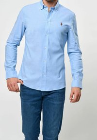 Man som bär en ljusblå skjorta med knappar och mörkblå jeans, stående med ena handen i fickan mot en enkel bakgrund.