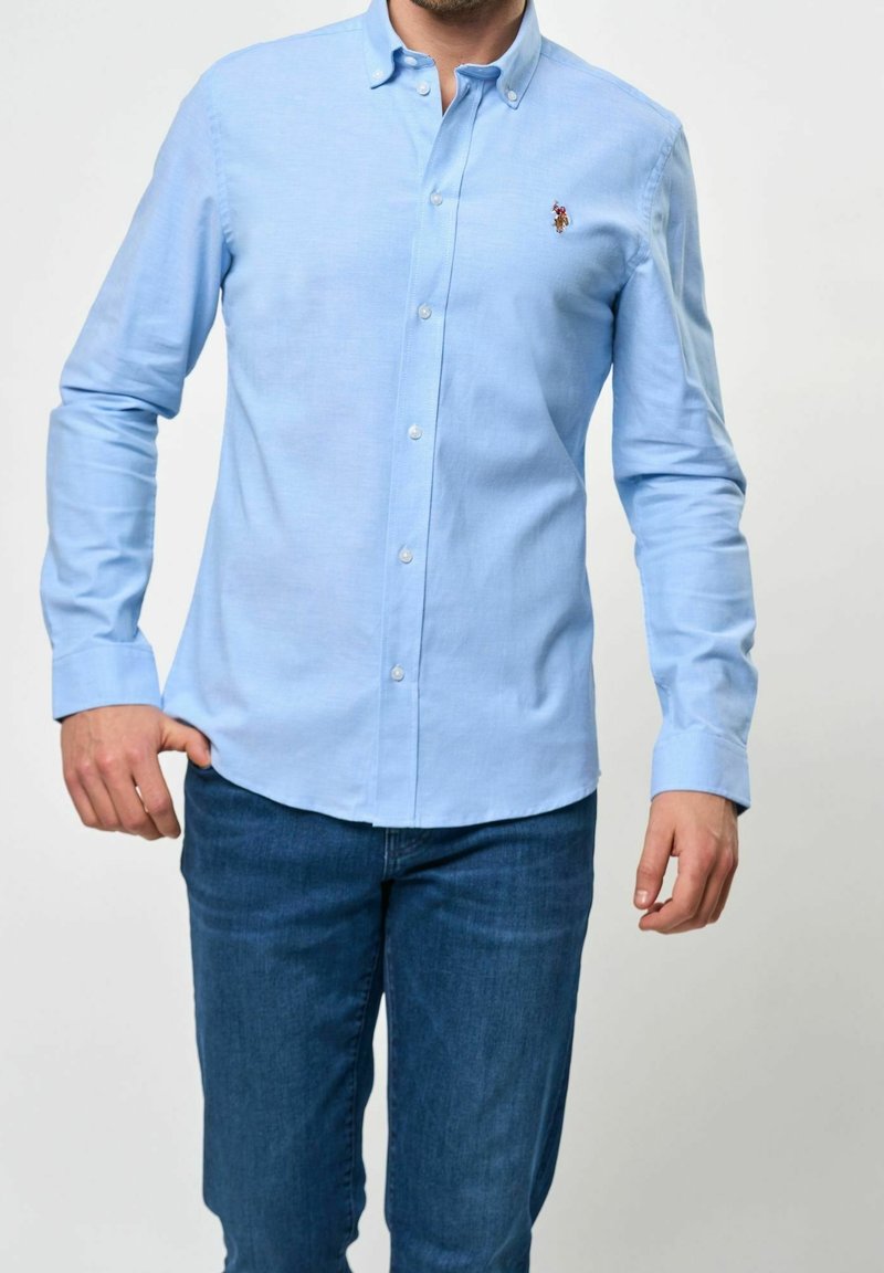 Man som bär en ljusblå skjorta med knappar och mörkblå jeans, stående med ena handen i fickan mot en enkel bakgrund.