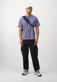 Lilla kortermet T-skjorte, svarte cargo-bukser med sidelommer, hvite joggesko, og en hvit caps, med en logo på skjorten og buksene.